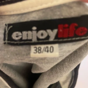 Grå t-shirt Enjoy Life v-ringad - Grå t-shirt från Enjoy Life med v-ringning och vita samt svarta detaljer vid hals och ärmslut. Tryck med texten 'ENJOY life' framtill. Skön passform och klassisk look, perfekt till jeans eller shorts.