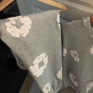 Denim tears byxor  - Grå mjukisbyxor med vita blommiga tryck över hela byxorna. De har resår i bensluten och midjan samt snörning för extra komfort. Perfekta för en chill och avslappnad stil.