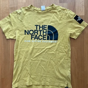 Gul t-shirt från The North Face - Gul t-shirt från The North Face med stor svart logga och text på bröstet samt patch med logga på ärmen. Klassisk rund hals och korta ärmar. Tillverkad i mjuk bomull, perfekt för en chill och sportig stil.