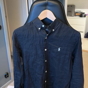Svart skjorta från Polo Ralph Lauren -  Svart linne skjorta från Polo Ralph Lauren i slim fit/S. Skjortan har klassisk krage med knappar, lång ärm och broderade loggan på bröstet. Tillverkad i bomull och passar perfekt till sommarn. Säljer pga att jag inte använder den längre. 