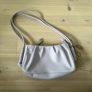 Lilac axelväska från Koan i skinnimitation - Snygg lilac axelväska från Koan i mjuk skinnimitation. Väskan har en stilren design med lätt rynkade detaljer, justerbar axelrem och guldfärgad dragkedja. Perfekt storlek för det viktigaste och med innerfack för extra organisering.