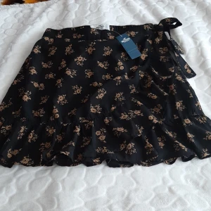 Svart blommig omlottkjol Hollister - Helt ny med prislappen på. Svart omlottkjol från Hollister med beige blommönster och volangkant. Kjolen har knyt i sidan och är i en luftig, lätt polyester. Ultra high-rise midja ger en snygg siluett. Perfekt för dig som gillar en feminin och trendig stil.