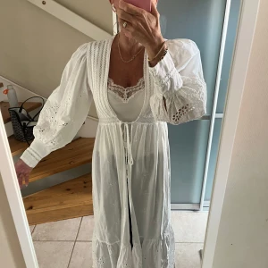 Vit långklänning med broderade detaljer - Superfin vit långklänning med broderade blommor och volang nertill. Klänningen har puffiga långärmar med spets och knappar vid ärmslut, samt knytband i midjan. Perfekt för sommarens alla tillfällen och ger en romantisk vibe.