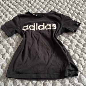 Svart Adidas t-shirt med logga - Svart t-shirt från Adidas med stor vit logga över bröstet och liten logotyp på ärmen. Klassisk rund hals och korta ärmar. Tillverkad i mjuk bomull, perfekt för sport eller chill. Enkel och stilren design som funkar till det mesta.