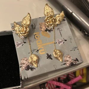 Guldiga örhängen med ros och löv från Ioaku - Unika örhängen i guldton från Ioaku med detaljerade rosor och löv. Hängande kedja med löv. Oanvände och box tillkommer. Nypris 700kr