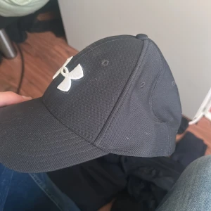 Svart keps från Under Armour - Svart keps från Under Armour med böjd skärm och klassisk passform. Kepsen har ett vitt broderat UA-logotyp framtill och är tillverkad i ett slitstarkt syntetmaterial. Perfekt för sportiga outfits eller när du vill hålla solen borta.