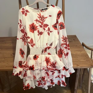 Vit blommig kortklänning Gina Tricot - Superfin vit kortklänning från Gina Tricot med röda blommor, volangkjol och smock i midjan. Långa ärmar med smock vid ärmslut och lätt transparent material ger en luftig känsla. Perfekt för sommarens alla tillfällen!