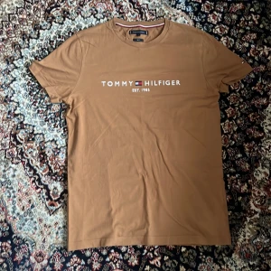 Brun Tommy Hilfiger t-shirt slim fit - Snygg brun t-shirt från Tommy Hilfiger i slim fit-modell. T-shirten har korta ärmar, rund halsringning och ett stort tryck med logga och texten 'Tommy Hilfiger Est. 1985' på bröstet. Tillverkad i mjuk bomull för skön känsla.