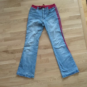 Blå bootcut jeans med röd snörning - Snygga ljusblå bootcut jeans med röd ribbad midja och snörning. Byxorna har röda ränder längs sidorn. Klassiska fickor fram och bak. Det är uppsprätta längs ner för att de skulle bli lite längre. 