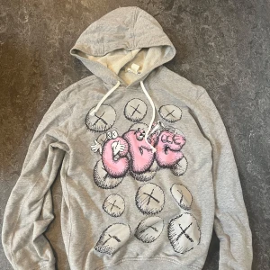 Grå hoodie Comme des Garçons x KAWS - Säljer en grå hoodie från Comme des Garçons med KAWS-print i rosa och vitt framtill. Tröjan har huva med snörning och är tillverkad i mjuk bomull. 