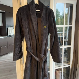 Morgonrock Lyle & Scott - Svart morgonrock från Lyle & Scott i mjuk frotté med klassisk omlottmodell och knytskärp i midjan. Liten gul logga på bröstet ger en clean och stilren look. Perfekt för chill dagar hemma eller efter duschen. Sparsamt använd, storlek medium! 