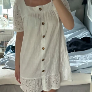 Vit kortklänning med broderade detaljer - Superfin vit kortklänning med broderade puffärmar och volangkant. Knäppning med stora knappar framtill och söt fyrkantig ringning. Perfekt för sommarens alla tillfällen! Passar S/M/L