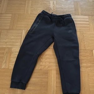 Svarta mjukisbyxor och flis från Nike - Svarta mjukisbyxor från Nike med dragsko i midjan och ribbade muddar vid bensluten. Byxorna har en slim passform och dragkedjeförsedda fickor på sidorna. Perfekta för chill eller träning, tillverkade i mjukt bomullsmaterial. Det ingår också en flis som är Nike tech samma med byxorna