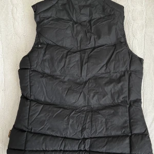 Svart dunväst från Nimbus XS - Svart dunväst från Nimbus i storlek XS. Västen har flera smarta innerfickor märkta för mobil och mp3, quiltad design och hög krage. Perfekt för lager-på-lager och har en clean, sportig look. Materialet är syntet och polyester.