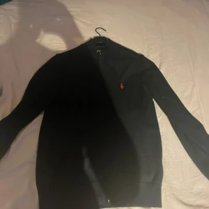 Svart kofta från Polo Ralph Lauren - Svart kofta från Polo Ralph Lauren med dragkedja framtill och klassisk röd logga broderad på bröstet. Långärmad modell i mjuk bomull, perfekt för lager-på-lager. Enkel och stilren design som funkar till det mesta.