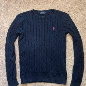 Mörkblå stickad tröja Polo Ralph Lauren - Snygg mörkblå kabelstickad tröja från Polo Ralph Lauren i bomull. Klassisk modell med rund halsringning, långa ärmar och rosa broderad logga på bröstet. Perfekt för dig som gillar stilrena och tidlösa plagg med en sportig touch. S passar XS