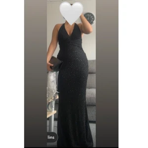 Svart glittrig långklänning med öppen rygg - Supersnygg svart långklänning med glittriga detaljer och djup v-ringning. Klänningen har halterneck och en öppen rygg som ger en riktigt elegant look. Sitter tight och framhäver figuren, perfekt för bal eller fest. Materialet känns lyxigt och skimrar snyggt i ljuset. Använt en gång bara. Nypris: 1900
