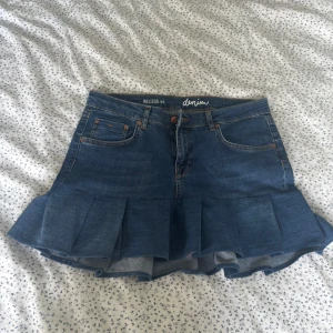 Jeanskjol med volang - Snygg blå jeanskjol med volang nertill och klassiska fickor fram och bak. Kjolen har en kort modell och stängs med knapp och dragkedja. Perfekt för dig som gillar denim och vill ha en cool twist på din outfit. Det står att den är storlek 44 men jag skulle säga att den passar en M/38.