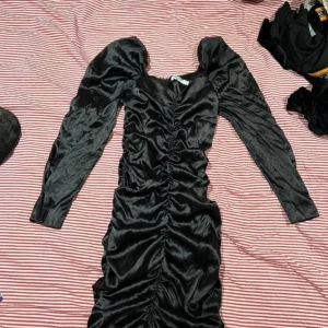 Svart klänning med rynkdetaljer - Snygg svart klänning i glansigt material med rynkdetaljer längs hela framsidan. Klänningen har långa ärmar och en figurnära passform som ger en elegant look.