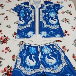 Blå sett med skjorta och shorts från Casablanca - Säljer ett sett från Casablanca med ett unikt mönster av vita svanar och blommor. Skjortan har en klassisk krage och knappar framtil. Perfekt för en stilren och iögonfallande look. 🌊🏖☀️🌴