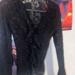 Svart spetsblus med volang - Elegant svart spetsblus med volangdetaljer framtill. Blusen har långa ärmar och ett transparent mönster som ger en sofistikerad look. Perfekt för en stilfull kväll.