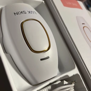 Lux Skin IPL Hårborttagningsenhet - Effektiv IPL-enhet från Lux Skin för hårborttagning. Elegant vit design med gulddetaljer. Kommer med strömkabel och användarmanual. Perfekt för smidig och smärtfri hårborttagning hemma.