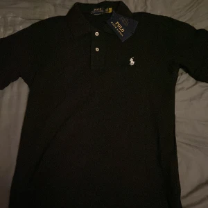 Svart pikétröja från Ralph Lauren - Säljer en klassisk svart pikétröja från Ralph Lauren med den ikoniska broderade loggan på bröstet. Tröjan har en slim fit passform och korta ärmar, perfekt för en stilren look. Två knappar vid kragen ger en snygg detalj. Passar utmärkt för både vardag och mer uppklädda tillfällen.