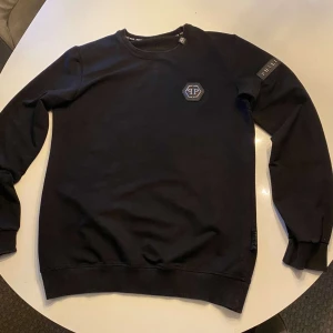 Svart tröja från Philipp Plein - Säljer en stilren svart tröja från Philipp Plein med ett diskret märke på bröstet. Tröjan har långa ärmar och rund halsringning. Perfekt för en casual look.