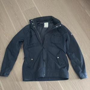 Mörkblå jacka från J.Lindeberg - Säljer en stilren mörkblå jacka från J.Lindeberg. Jackan har långa ärmar och flera praktiska fickor med knappar. Den är utrustad med både dragkedja och knappar framtill för extra skydd mot väder. Perfekt för vår och höst!