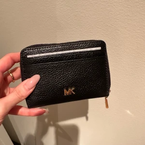 Svart plånbok från Michael Kors - Snygg svart plånbok i skinn från Michael Kors med guldfärgad dragkedja och logotyp. Perfekt för att hålla ordning på kort och kontanter. Kompakt och stilren design som passar i alla väskor.