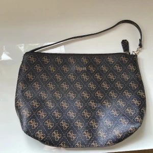 Svart och brun axelväska från Guess - Snygg axelväska från Guess med ett klassiskt monogrammönster i svart och brunt. Väskan har en dragkedja upptill och en justerbar axelrem. Perfekt för att bära dina nödvändigheter med stil.