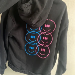 Svart hoodie med smiley-motiv - Säljer en svart hoodie med färgglada smiley-ansikten i blått och rosa på ryggen. Hoodien har en klassisk design med huva och långa ärmar. Perfekt för en avslappnad stil.