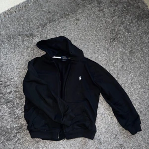 Svart huvtröja från Polo Ralph Lauren - Säljer en svart huvtröja från Polo Ralph Lauren med dragkedja och broderad logga på bröstet. Tröjan har långa ärmar och en praktisk huva. Perfekt för en avslappnad stil.