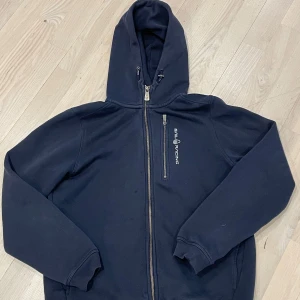 Svart hoodie från Sail Racing - Säljer en svart hoodie från Sail Racing med dragkedja och huva. Tröjan har en stilren design med logotypen broderad på bröstet. Perfekt för en avslappnad stil.