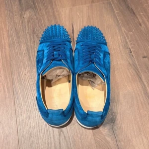 Blå sneakers med nitar - Snygga blå sneakers i mocka med nitar på tån från Christian Louboutin. Skorna har en ikonisk röd sula och snörning framtill. Perfekta för att ge din outfit en edgy touch. byten tillgänglig 💫🙌