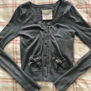 Grå kofta från Abercrombie & Fitch - Säljer en grå kofta från Abercrombie & Fitch med långa ärmar och söta rosetter på fickorna, en av sömmen på fickorna gått av (se bild) Storlek S/XS, priset går att diskutera !