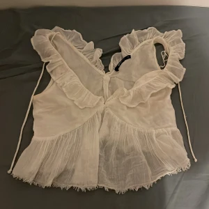 Vit sommar blus från Gina Tricot.  - Säljer en söt vit blus från Gina Tricot med volanger och knytband. Blusen har en luftig design med ärmlös stil och en romantisk touch. Perfekt för varma dagar eller sommarens fester.