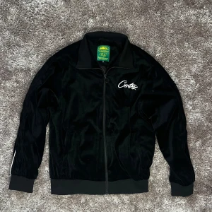 Corteiz zip up - Snygg svart jacka från Corteiz med dragkedja och vit logga på bröstet och ryggen. Jackan har långa ärmar och en stilren design med vita detaljer längs ärmarna. Perfekt för en cool och avslappnad stil.