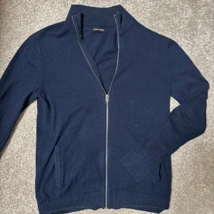  Jack & Jones fullzip - Säljer en stilren mörkblå kofta från Jack & Jones med dragkedja framtill. Koftan har långa ärmar och ribbade muddar vid ärmslut och nederkant. Perfekt för en avslappnad look.