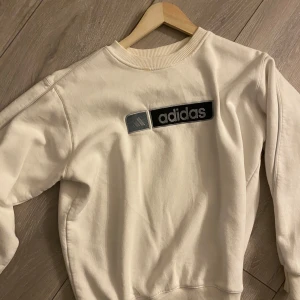 Vit sweatshirt från Adidas - Säljer en stilren vit sweatshirt från Adidas med deras klassiska logga på bröstet. Tröjan har långa ärmar och en rund halsringning med ribbade kanter. Perfekt för en avslappnad look. Liten defekt vid kragen vet ej om den går att få bort