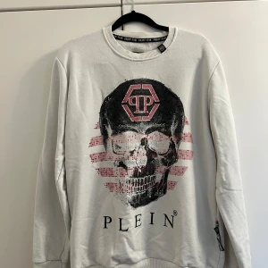 Vit tröja med dödskalle från Philipp Plein - Säljer en cool vit tröja från Philipp Plein med ett stort dödskalletryck och logotyp i svart och rött. Tröjan har långa ärmar och en rund halsringning. Perfekt för dig som gillar en edgy stil!