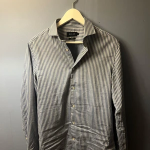 Skjorta från Riley - Snygg randig skjorta från Riley i blått och vitt. Skjortan har en slim fit passform och är långärmad med knappar framtill. Perfekt för en stilren look. Priset kan diskuteras vid snabb affär! denna skjorta är bara använd nåra fåtal gånger!