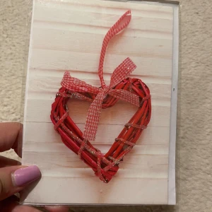 Kort med rött hjärta och rosett - Charmigt kort med ett rött hjärta dekorerat med en glittrig rosett och band. Perfekt för att skicka en kärleksfull hälsning eller som en fin detalj till en present. Kortet har en rustik träbakgrund som ger en hemtrevlig känsla. Ett kurvet följer med. 