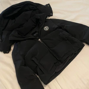 Svart pufferjacka från Trapstar - Säljer en svart pufferjacka från Trapstar med dragkedja och huva. Jackan har en cool logga på bröstet och text på ryggen. Perfekt för kyliga dagar när du vill hålla stilen. 🖤