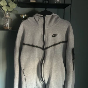 Grå Nike tech fleece från Nike - Säljer en grå huvtröja från Nike med dragkedja och svart logga på bröstet. Tröjan har långa ärmar och en praktisk ficka på ärmen. Perfekt för en avslappnad stil.