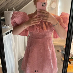 Rosa klänning med volangdetaljer - Charmig rosa klänning med volangdetaljer och offshoulder-design. Den har en texturerad yta som ger en unik look. Perfekt för speciella tillfällen!🩷 aldrig använd!! Står inget märke eller storlek då jag köpte den i en liten butik på Öland! Men sitter som en XS