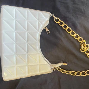Vit handväska  - Snygg vit handväska med quiltat mönster och en cool guldfärgad kedja som axelrem. Väskan har en praktisk dragkedja upptill för säker förvaring. Perfekt för att ge din outfit en trendig touch.
