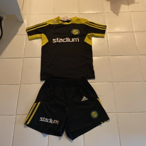 Svart och gul fotbollströja från Adidas - Snygg svart och gul handbollstjöja från Adidas med korta ärmar och klubbmärket för IK Sävehof. Tröjan har gula detaljer och Stadium-logga på framsidan. Perfekt för unga handbolssfans!