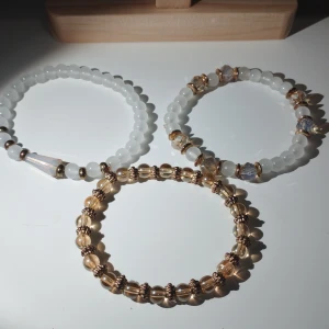 Set med tre armband i pärlor - Snyggt set med tre armband i olika stilar. Ett armband har vita pärlor med en konformad detalj, ett annat har vita och guldiga pärlor, och det tredje har genomskinliga och guldiga pärlor. Perfekt för att mixa och matcha! Diametern är 6 cm, omkretsen är cirka 16 cm.