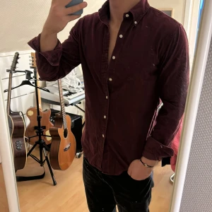 Vinröd manchester skjorta från Ralph Lauren - Snygg vinröd manchester skjorta från Ralph Lauren med klassisk krage och knappar framtill. Skjortan har en broderad logga i blått på bröstet och långa ärmar. Perfekt för en stilren look!